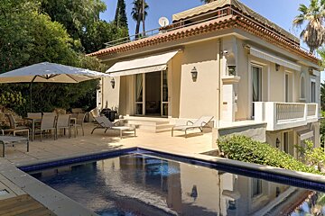 A cream-colored villa with a tiled roof overlooks a sparkling blue swimming pool and a sunny patio with outdoor dining and lounge chairs.