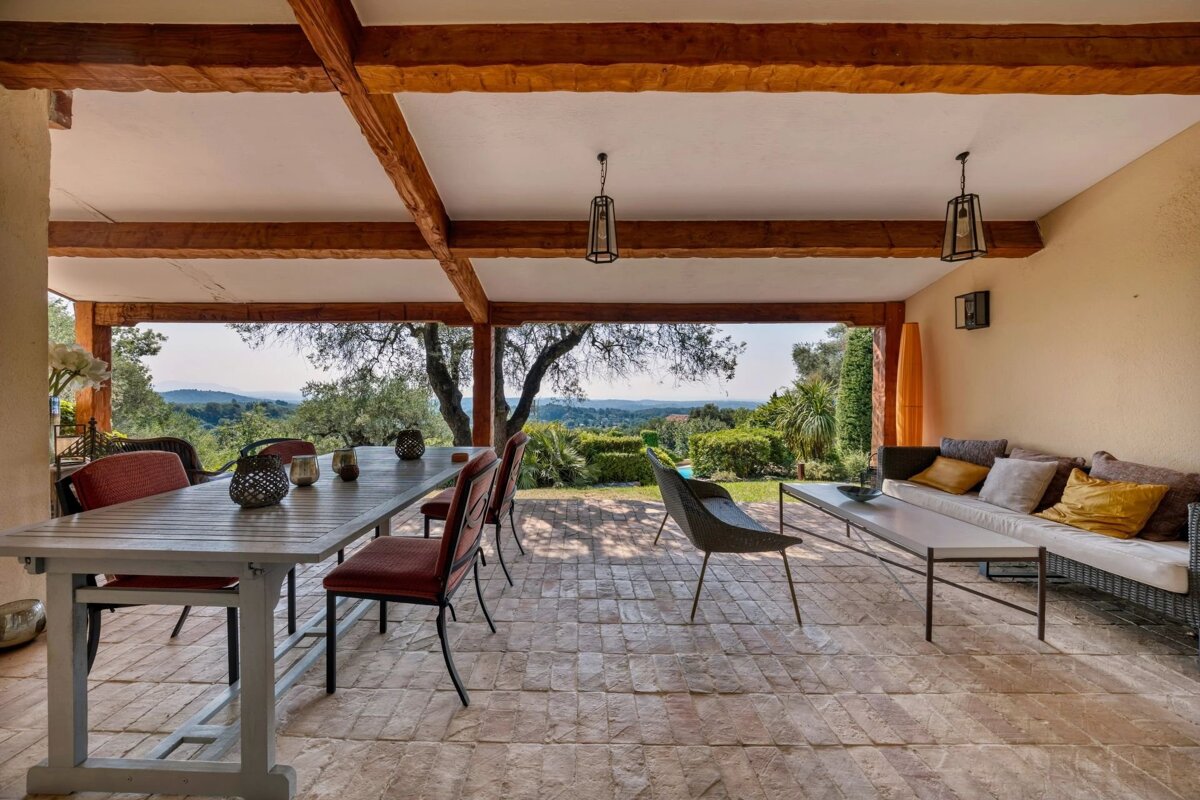 Covered patio with dining and lounge areas, featuring exposed wooden beams and scenic landscape views.