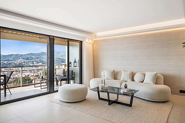 A modern, light-toned living room with a cream sofa, glass coffee table, and large sliding doors opening to a balcony with city and mountain views.
