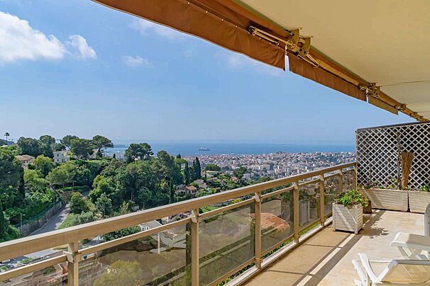 Balcony with an awning offering a stunning panoramic view of a coastal city, rolling green hills, and the calm blue sea.