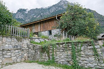 A rustic stone and wood house, framed by stone walls and lush greenery, sits below cloudy mountains with misty peaks.