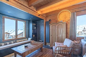 Cozy chalet interior with wooden walls, a window overlooking snowy mountains, a rustic coffee table, and an armchair.