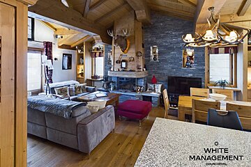 Cozy chalet living room with exposed wood beams, stone fireplace, moose head, comfortable sofas, and a dining area, exuding a rustic charm.