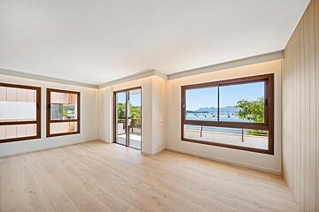 Bright, empty room with light wood floors, large windows, and a balcony door. One window overlooks a harbor with boats and mountains.