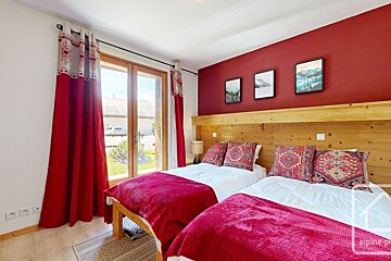 Cozy twin bedroom with red blankets, patterned cushions, and a vibrant red accent wall. Features a wooden headboard and a large window.