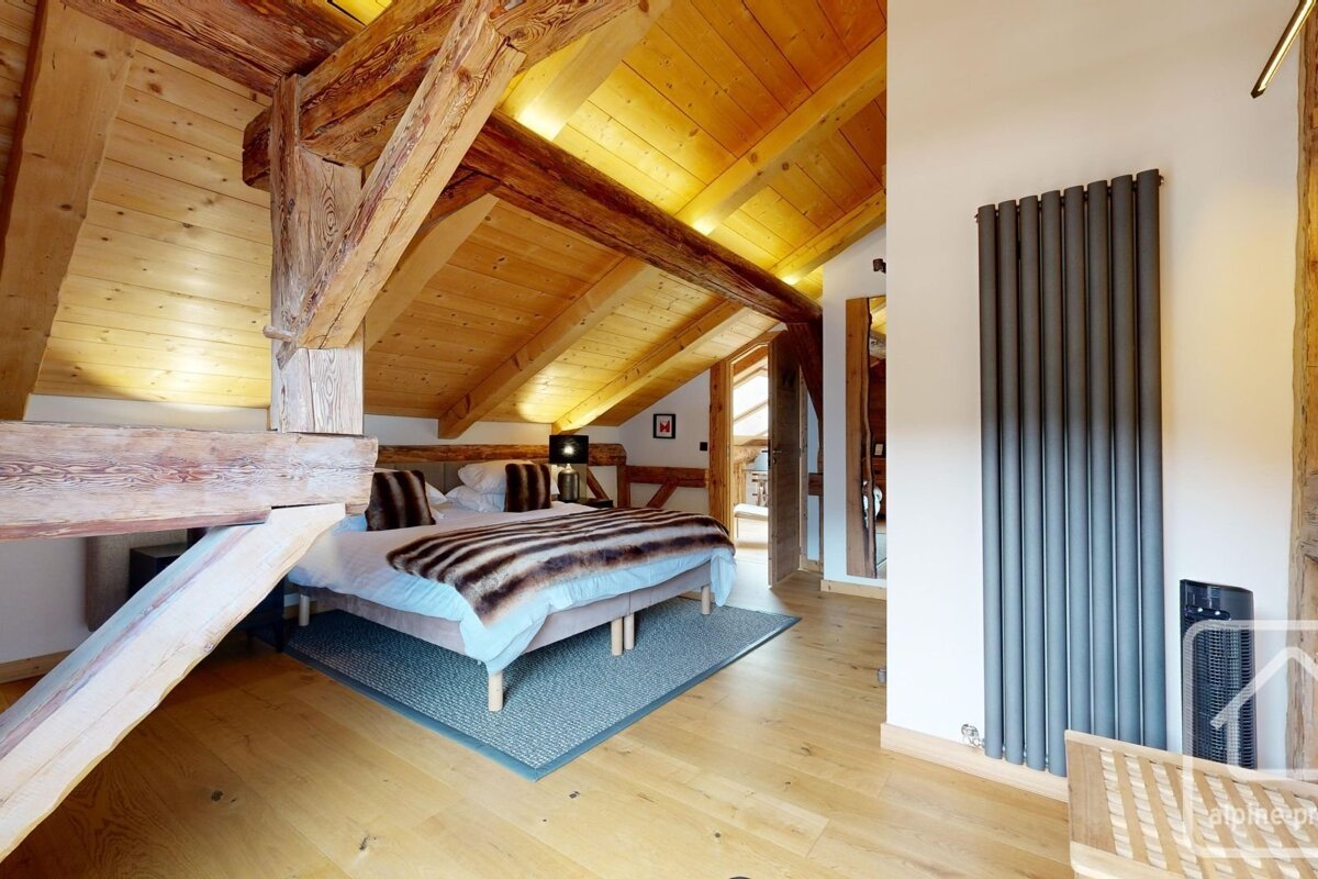 Rustic attic bedroom featuring prominent wooden beams, a sloping ceiling, and a cozy bed with a fur throw, beside a sleek modern radiator.