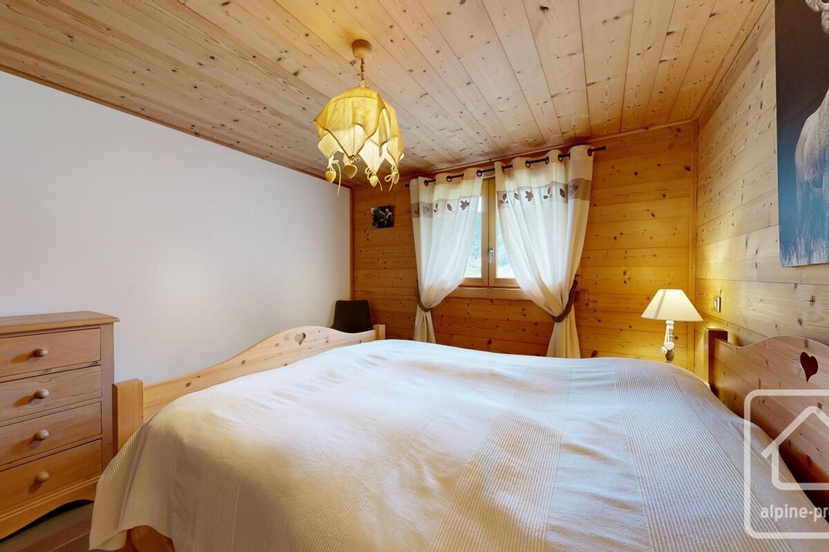 A charming cabin-style bedroom featuring wooden paneled walls and ceiling, a large bed with heart-carved headboards, a window, dresser, and unique chandelier.