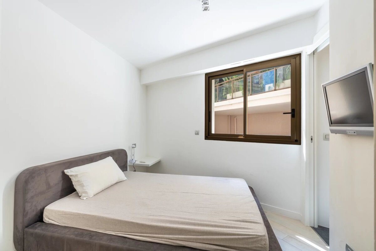 A minimalist bedroom with white walls, a grey upholstered bed, a window with a brown frame, and a wall-mounted TV.
