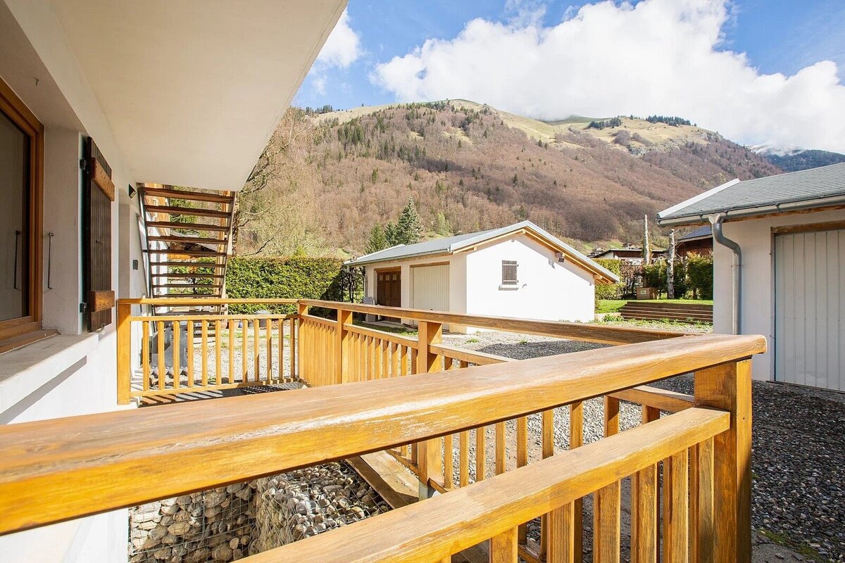 A wooden balcony or terrace offers a view of white buildings, a mountain covered in trees, and a partly cloudy sky. A gabion wall is visible below.