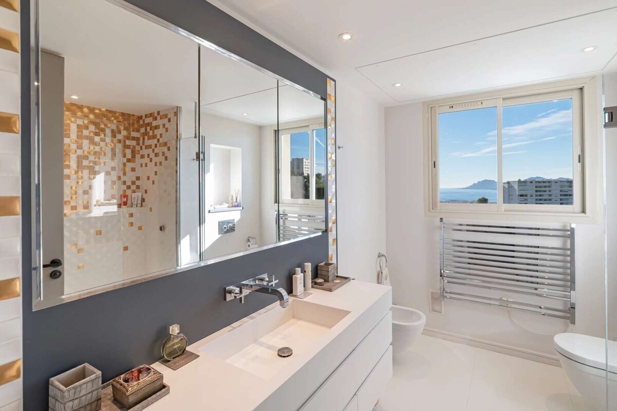 A modern, bright bathroom featuring a large mirror over a double vanity, a white and gold tiled shower, and a window with a scenic sea view.