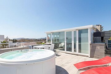 A modern rooftop terrace with a large white hot tub, red lounge chairs, outdoor dining area, and glass sunroom under a clear blue sky.