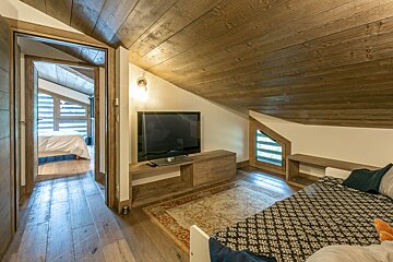 Cozy chalet-style room with a slanted wooden ceiling, a TV, a patterned rug, and a doorway opening to another bedroom.