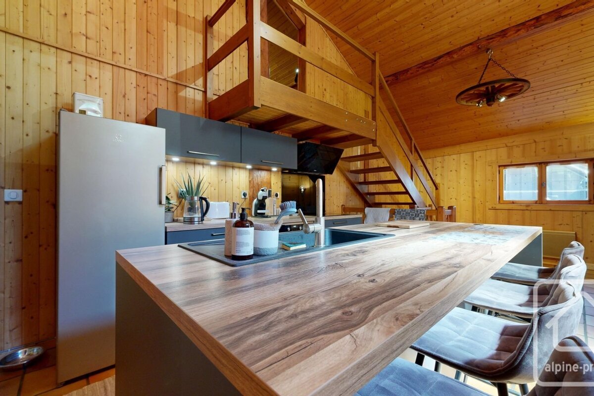 Cozy wooden cabin kitchen with a modern island, fridge, and a loft overhead. Natural wood walls and ceiling create a warm ambiance.