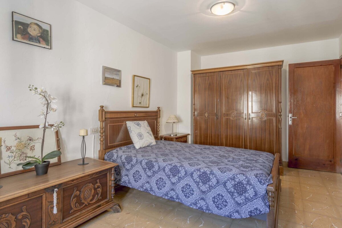 A simple bedroom featuring a single bed with blue patterned bedding, traditional wooden furniture (wardrobe, chest), white walls, and a tiled floor.