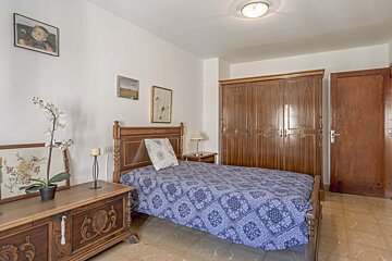A simple bedroom featuring a single bed with blue patterned bedding, traditional wooden furniture (wardrobe, chest), white walls, and a tiled floor.