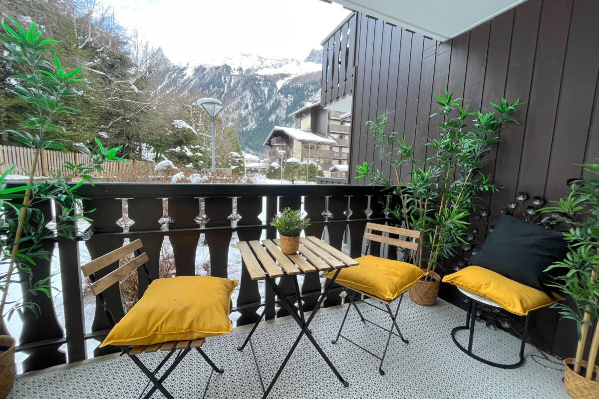 Cozy winter balcony with a small table, chairs, yellow cushions, and plants, offering a scenic view of snow-covered mountains and pine trees.