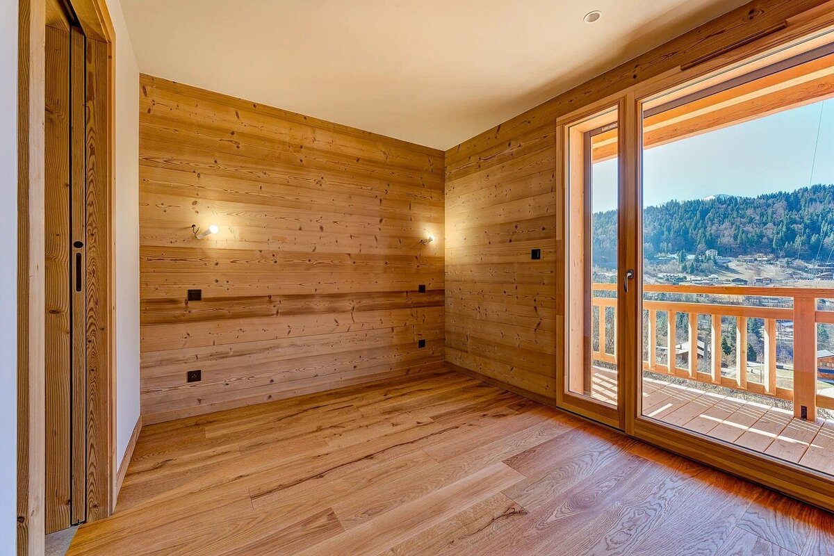 Empty wood-paneled room with a large glass door opening to a sunny balcony overlooking a scenic mountain landscape.