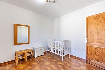 A simple room with white walls, brown tile flooring, a white baby crib, two wooden stools, a hanging mirror, and a wooden door.