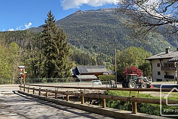 A sunny village scene in the mountains, featuring a bridge, houses, and a tractor, set against a large forested peak under a blue sky.
