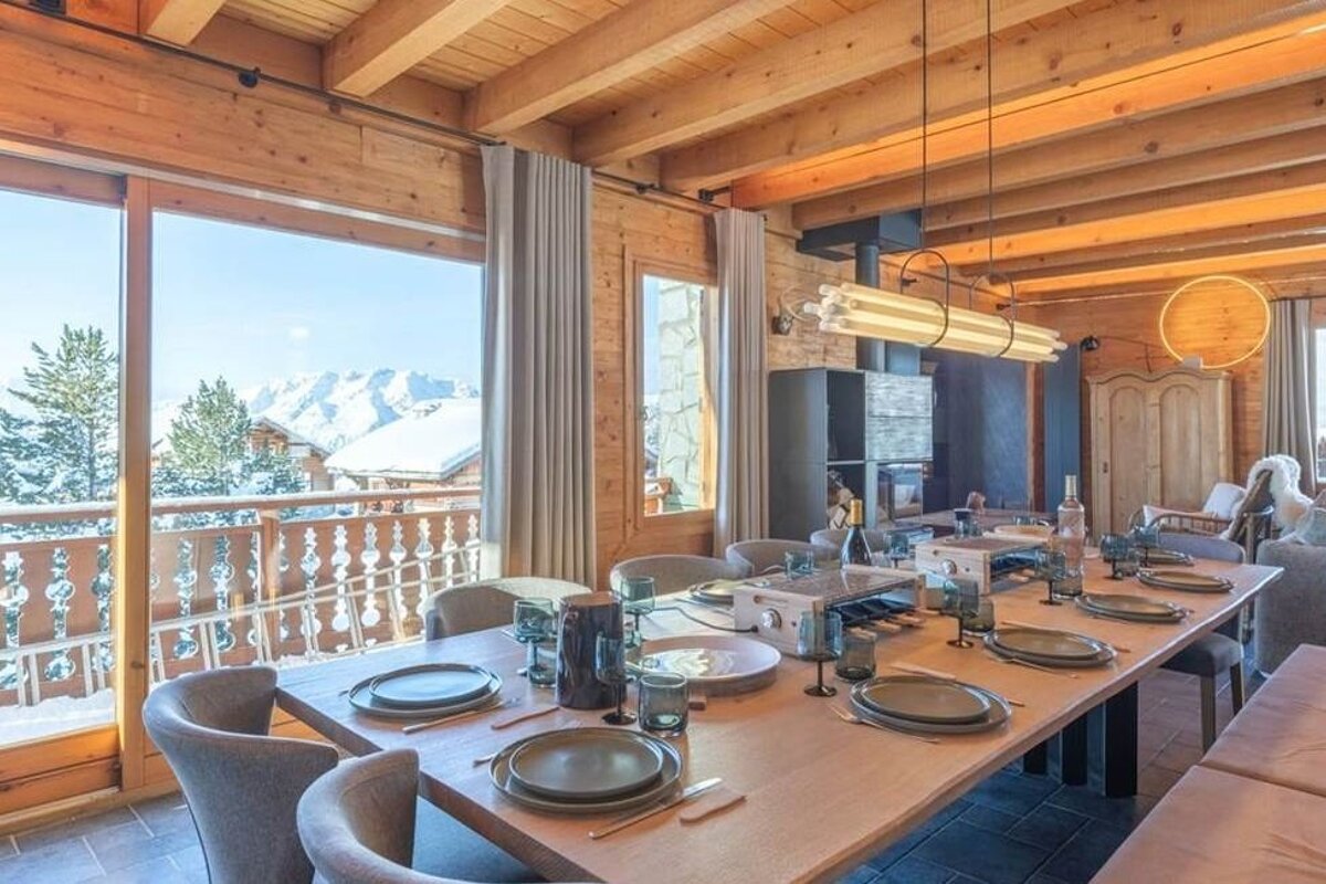 A long dining table set in a cozy wooden chalet with large windows offering a view of snow-covered mountains.