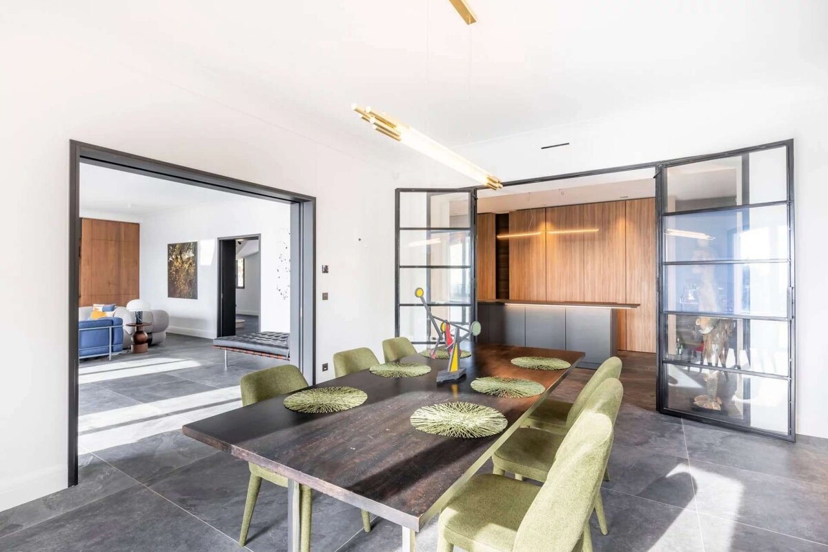 Bright, modern dining room with dark wood table, olive chairs, and steel-framed glass doors leading to a contemporary kitchen and living space.