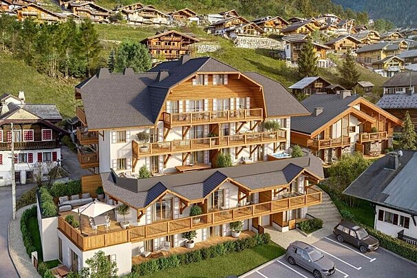 A large, modern chalet with balconies dominates a hillside village filled with traditional wooden houses, surrounded by green trees and mountains.