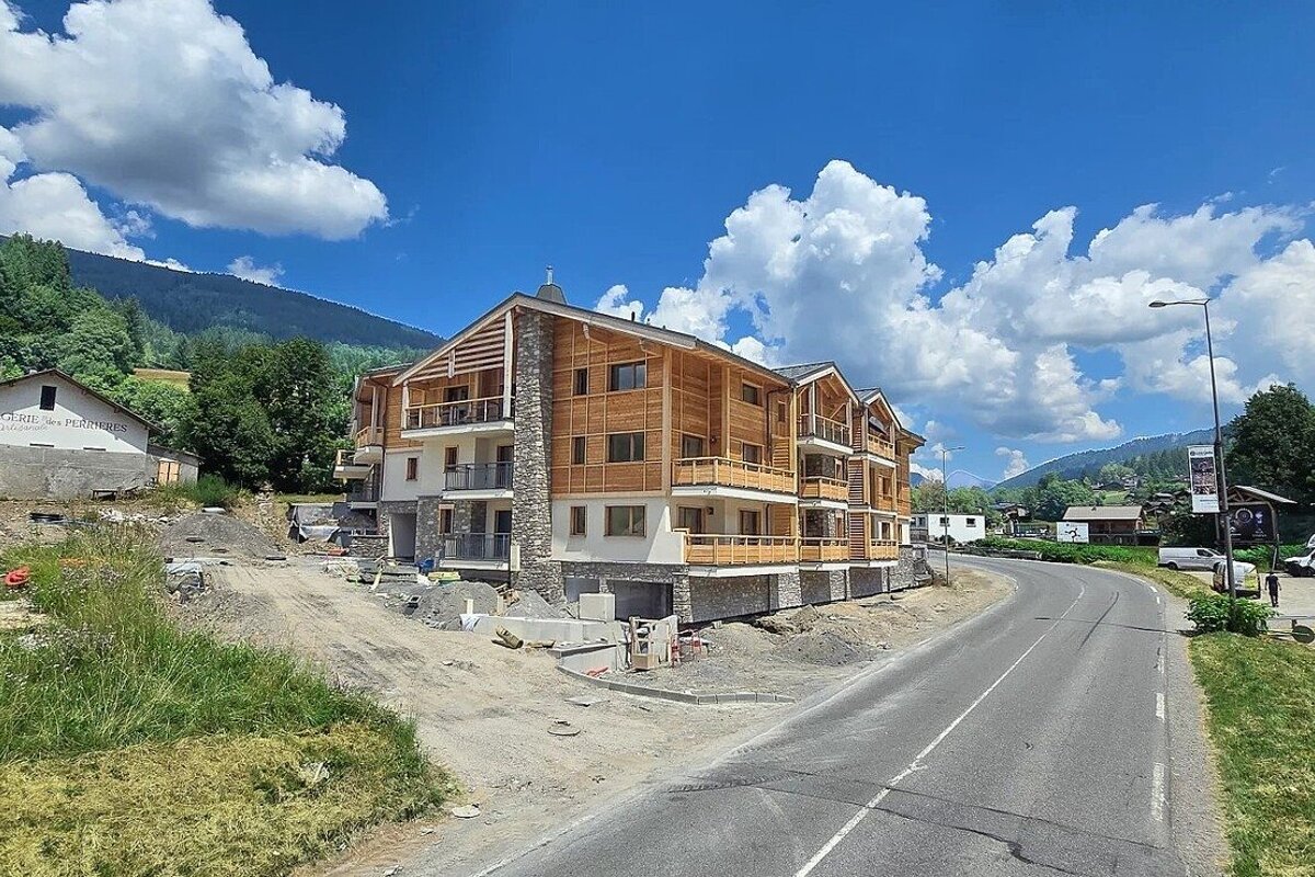 A new wood and stone chalet-style building under construction next to a road, with green mountains and a blue, cloudy sky in the background.