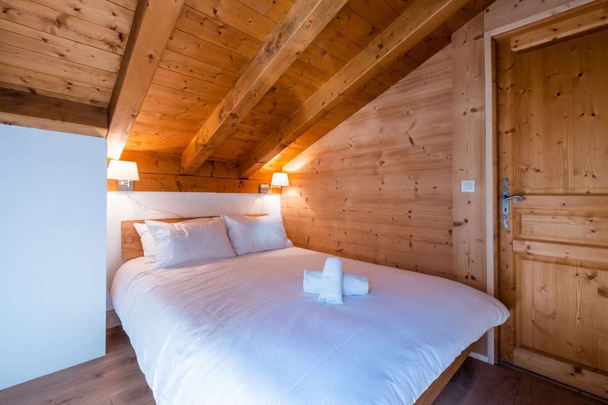 A cozy, wood-paneled attic bedroom features a white bed with pillows and neatly folded towels, illuminated by two wall lamps. A wooden door is on the right.