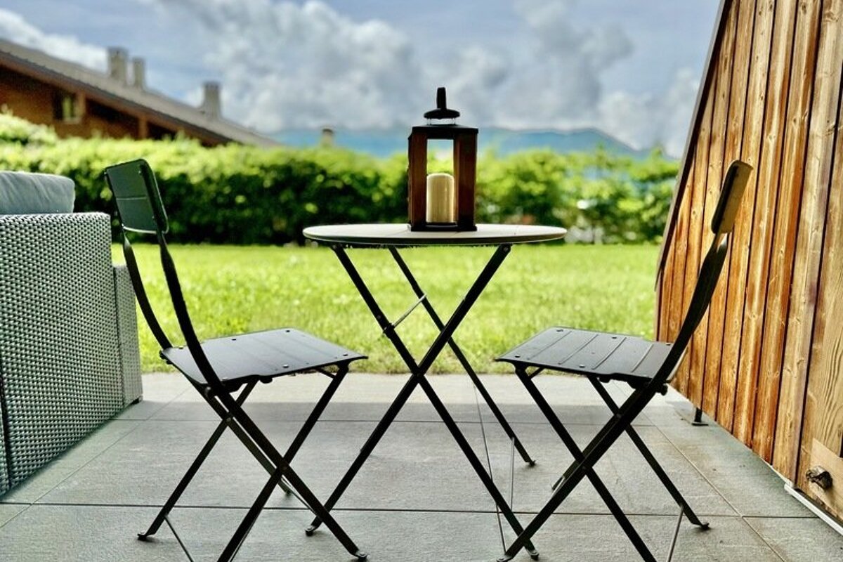 Two folding chairs and a small table with a lantern on a patio, overlooking a lush green lawn and distant mountains.