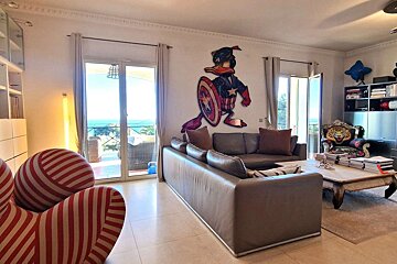A bright living room with an ocean-view balcony. Features a Captain America Donald Duck wall art, a brown sofa, a red striped chair, and a tiger chair.
