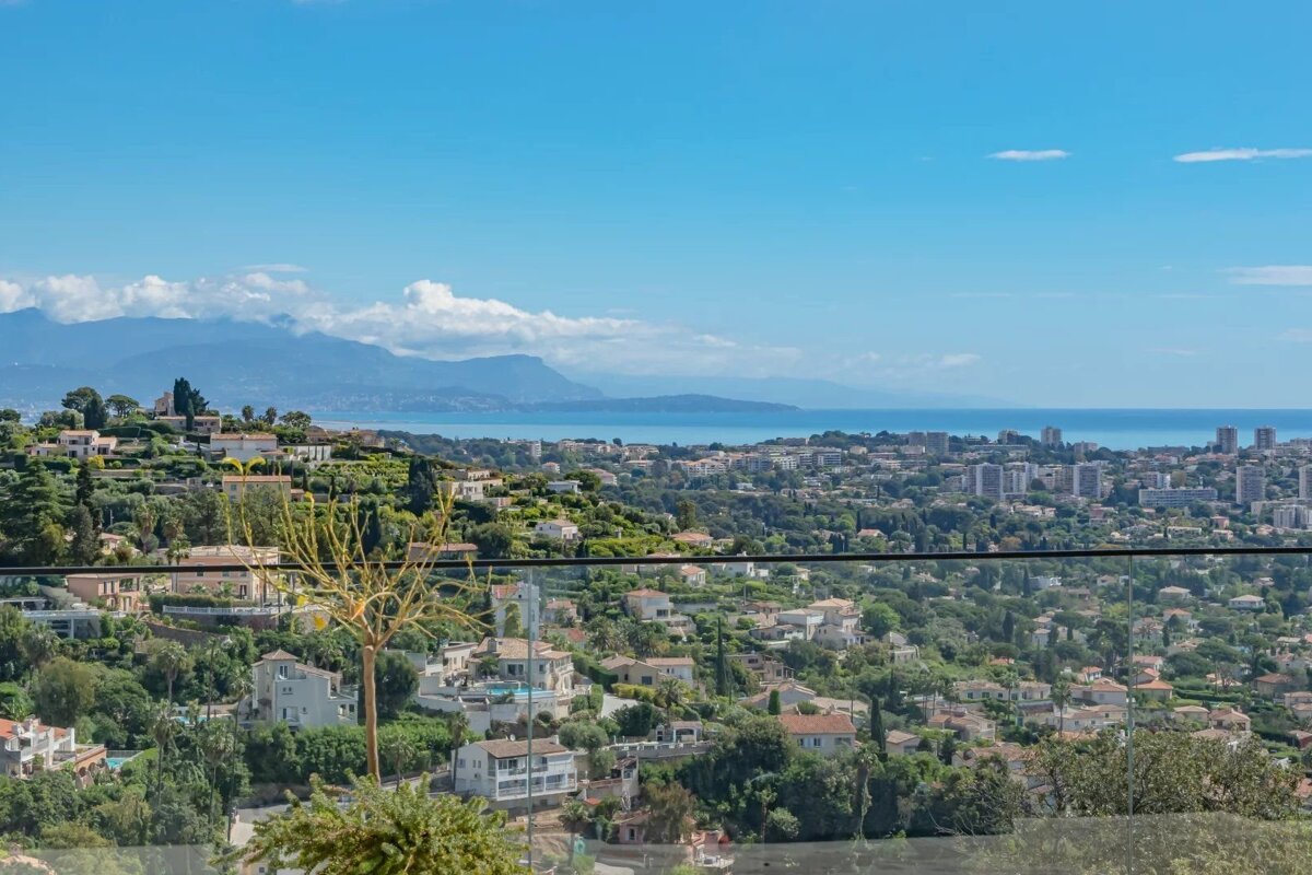 Panoramic view of a coastal city built into green hills, with distant mountains, the blue sea, and clear skies.