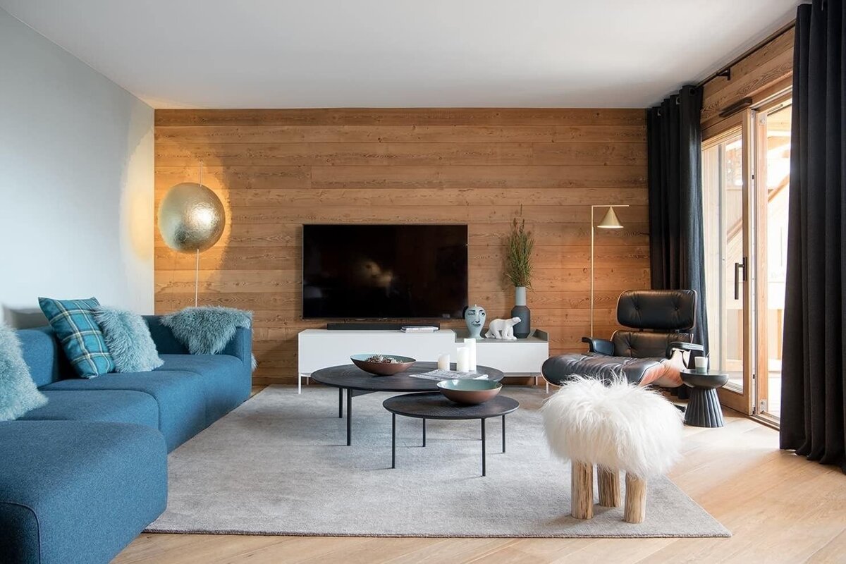 A cozy modern living room with a blue sofa, wood-paneled TV wall, black lounge chair, fluffy stool, and contemporary decor.