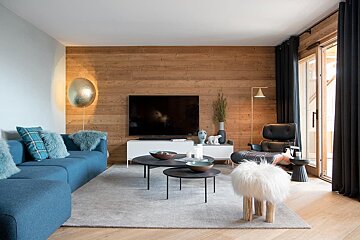 A cozy modern living room with a blue sofa, wood-paneled TV wall, black lounge chair, fluffy stool, and contemporary decor.