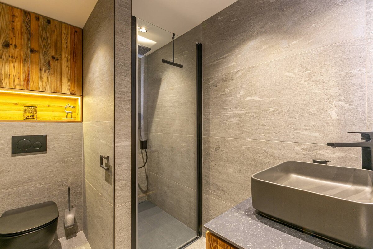 Sleek modern bathroom with gray tiles, black toilet, shower, and vessel sink. Warmly lit wooden shelf adds a rustic touch.