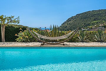 A hammock stands poolside with tropical plants and a hillside view under a clear blue sky.