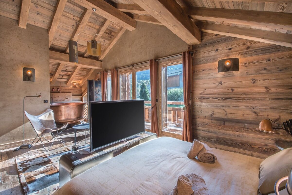 Luxurious chalet bedroom with exposed wooden beams, a large bed, TV, copper bathtub, and floor-to-ceiling windows with a scenic view.