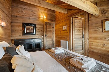 Rustic chalet bedroom featuring wood-paneled walls and ceilings, a bed with folded towels, and a dark wooden dresser with a mirror.