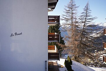 Alpine building "Le Rouet" with wooden balconies, overlooking a snowy mountain valley with tall pine trees under a clear blue sky.