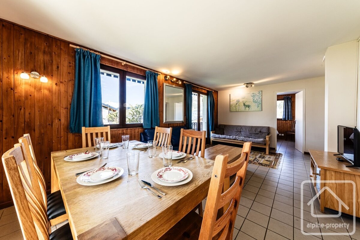 Bright, rustic dining & living area with wood-paneled walls, a large table set for guests, blue curtains, and a sofa in the background.