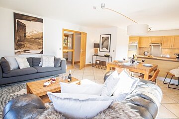 Bright open-plan living room with comfortable sofas, a wooden coffee table, dining area, and a kitchen.