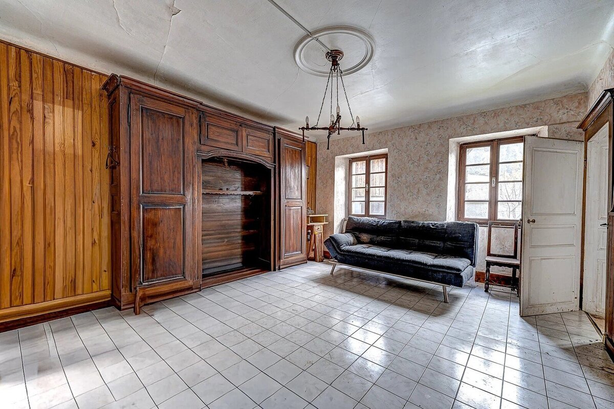 A vintage room with a large dark wood armoire, black sofa, tiled floor, and two windows. A chandelier hangs from the cracked ceiling.