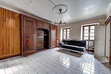 A vintage room with a large dark wood armoire, black sofa, tiled floor, and two windows. A chandelier hangs from the cracked ceiling.