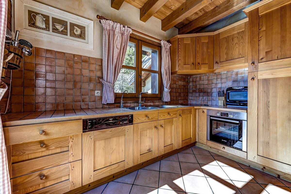 A rustic kitchen with light wood cabinets, terracotta tiles, exposed beams, and a window with gingham curtains. Features an oven, dishwasher, and sink.