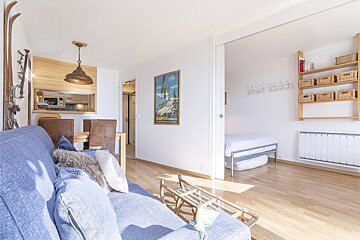 Bright, modern ski apartment with a blue sofa, dining area, kitchen pass-through, and a bedroom. Features skis, a sled, and a ski poster.