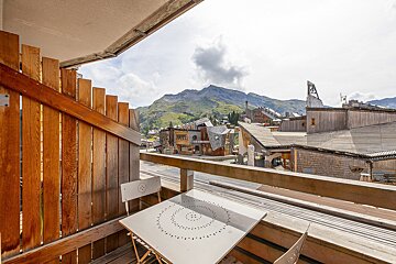 A wooden balcony with a white table and chairs overlooks a charming mountain village with distinctive buildings and lush green mountains under a cloudy sky.