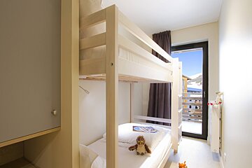 A bright bedroom features light wooden bunk beds with a stuffed owl, and a large window overlooks a snowy mountain landscape.