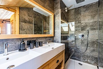 Modern rustic bathroom featuring grey stone tiles, a white double sink vanity, a large wood-framed mirror, and a glass shower.