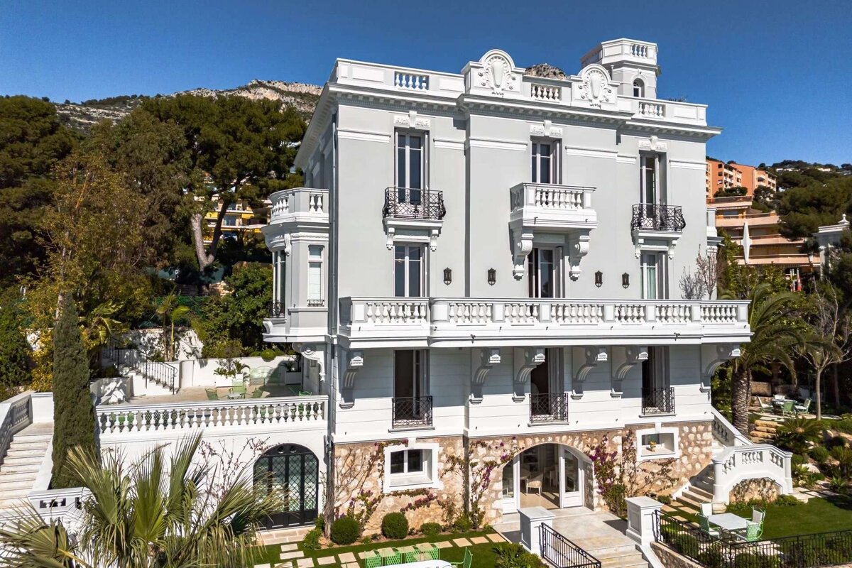 A stately light gray villa with ornate balconies and balustrades, surrounded by terraced gardens and lush hills under a clear blue sky.
