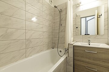 A bright, modern bathroom featuring a white tiled tub/shower, sink with wooden vanity, and mirror.