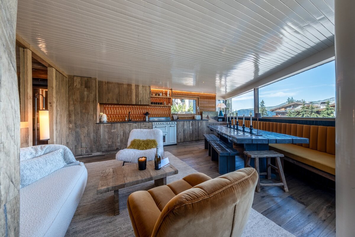 Modern chalet interior featuring a wood-paneled kitchen, cozy living area, and a long dining table by a large window with mountain views.